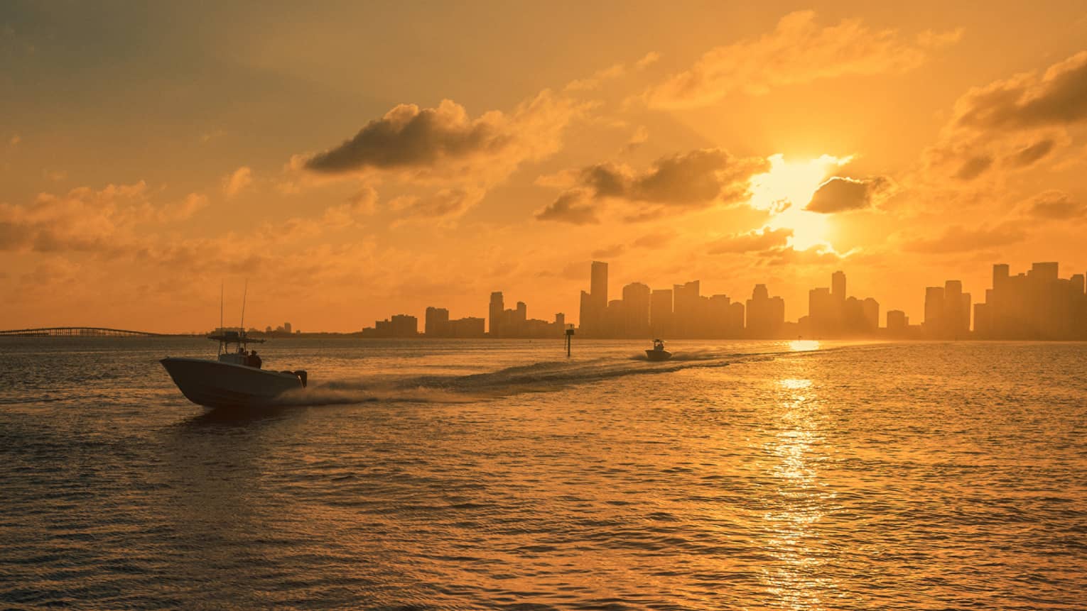 A boat cuts through ocean water in golden sunset light with a city skyline in the background
