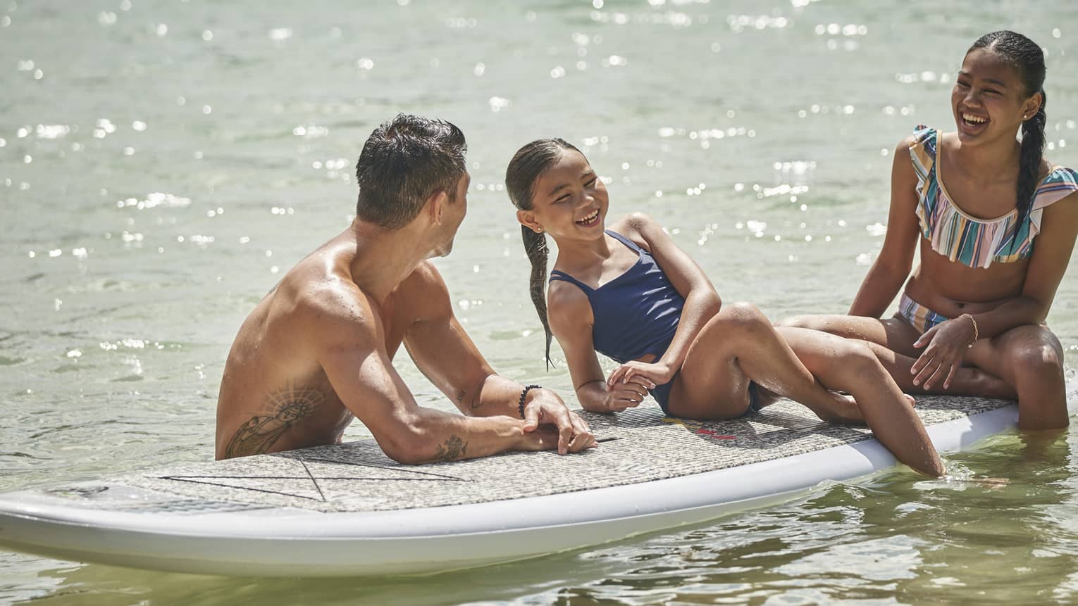 Two children sit on a stand-up paddleboard smiling as an instructor looks toward them holding the board still in the water