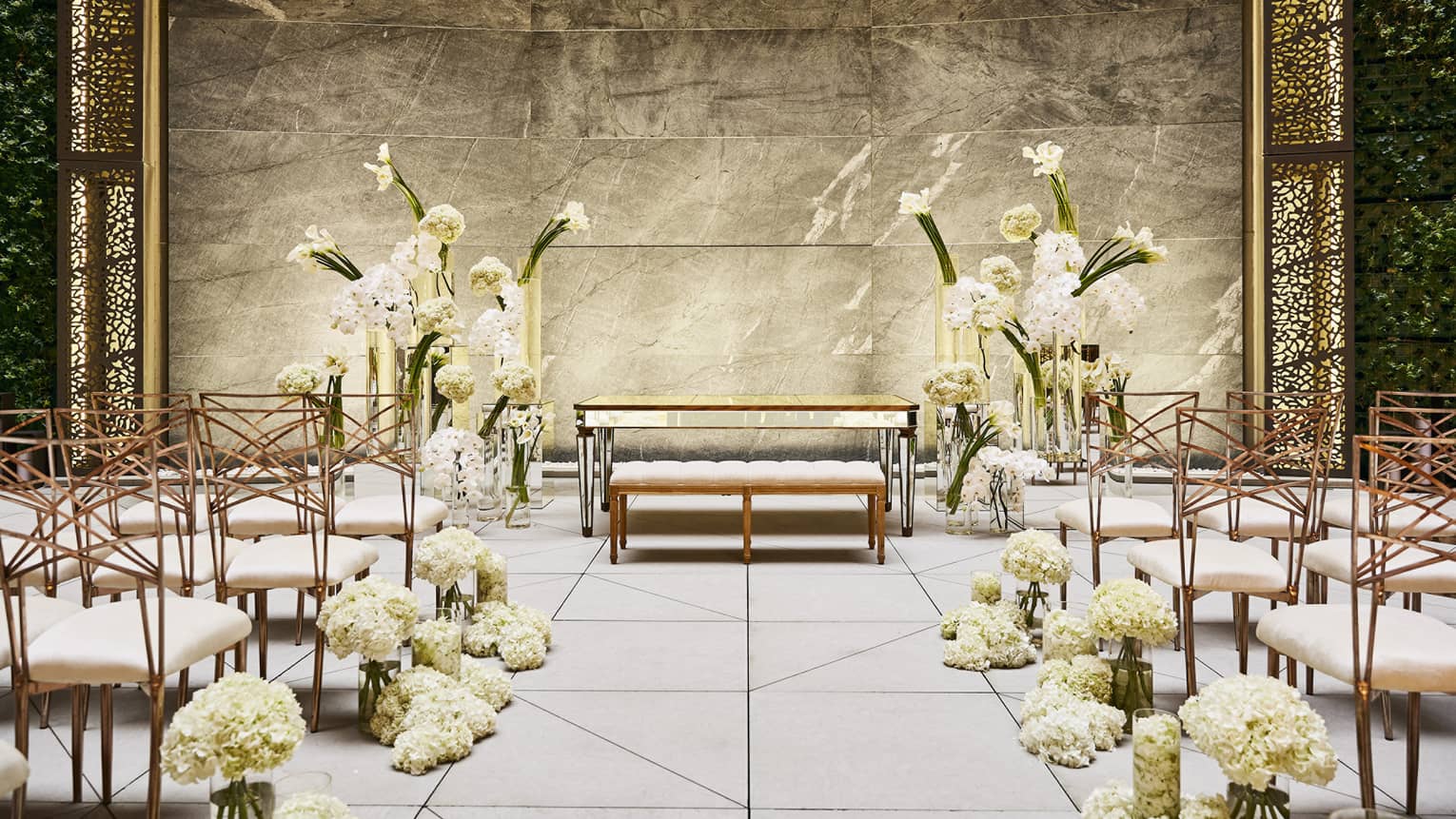 An elegant indoor wedding ceremony setup with white floral arrangements, modern chairs and a marble backdrop, featuring intricate gold latticework.