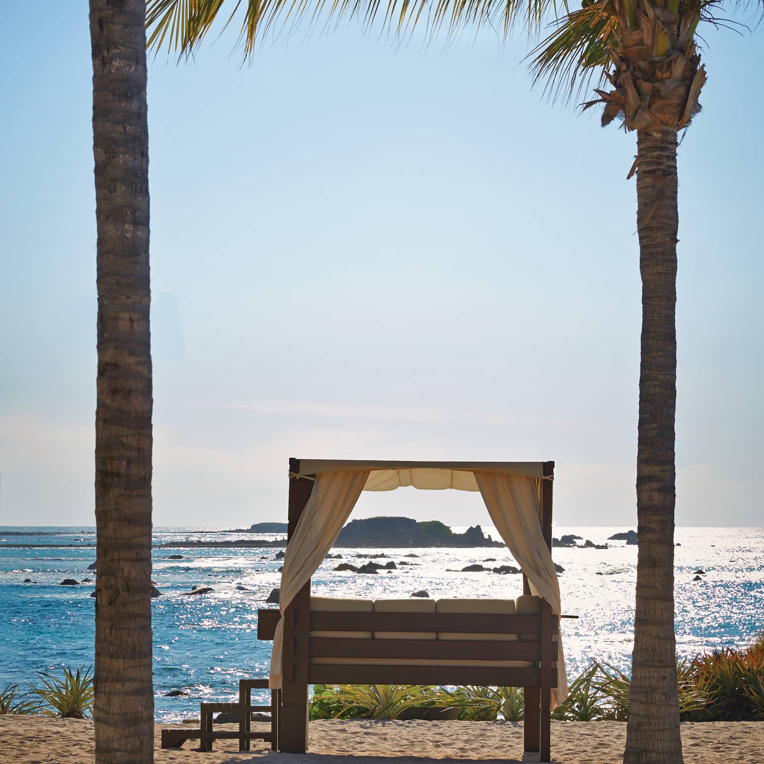 Steps lead up to a curtained daybed flanked by palm trees overlooking rocky islands in a sea of glimmering turquoise.