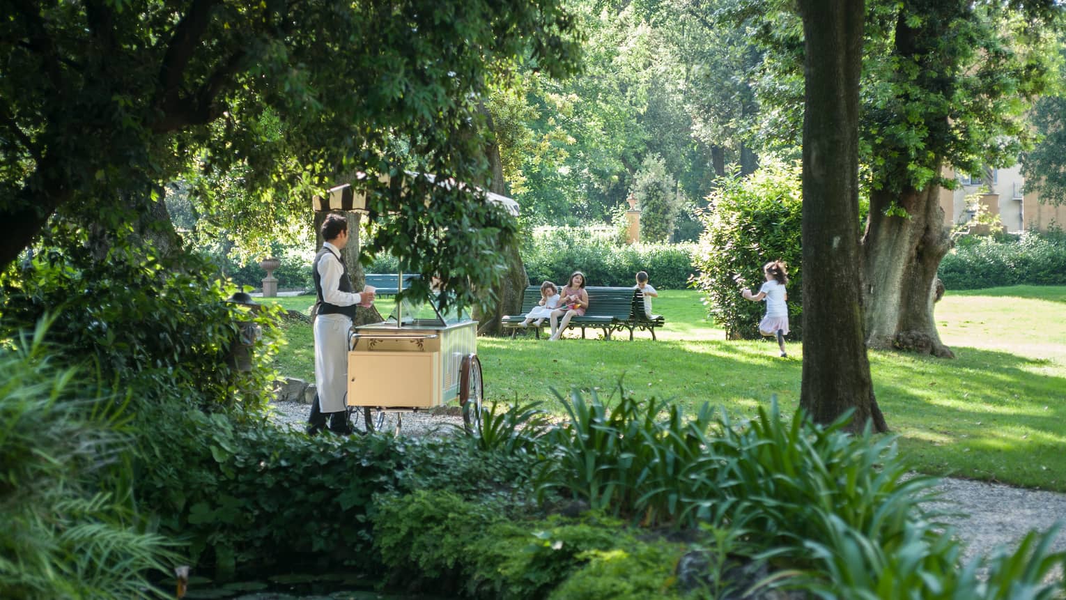 Man wearing apron stands behind ice cream cart, children sit, play on garden benches