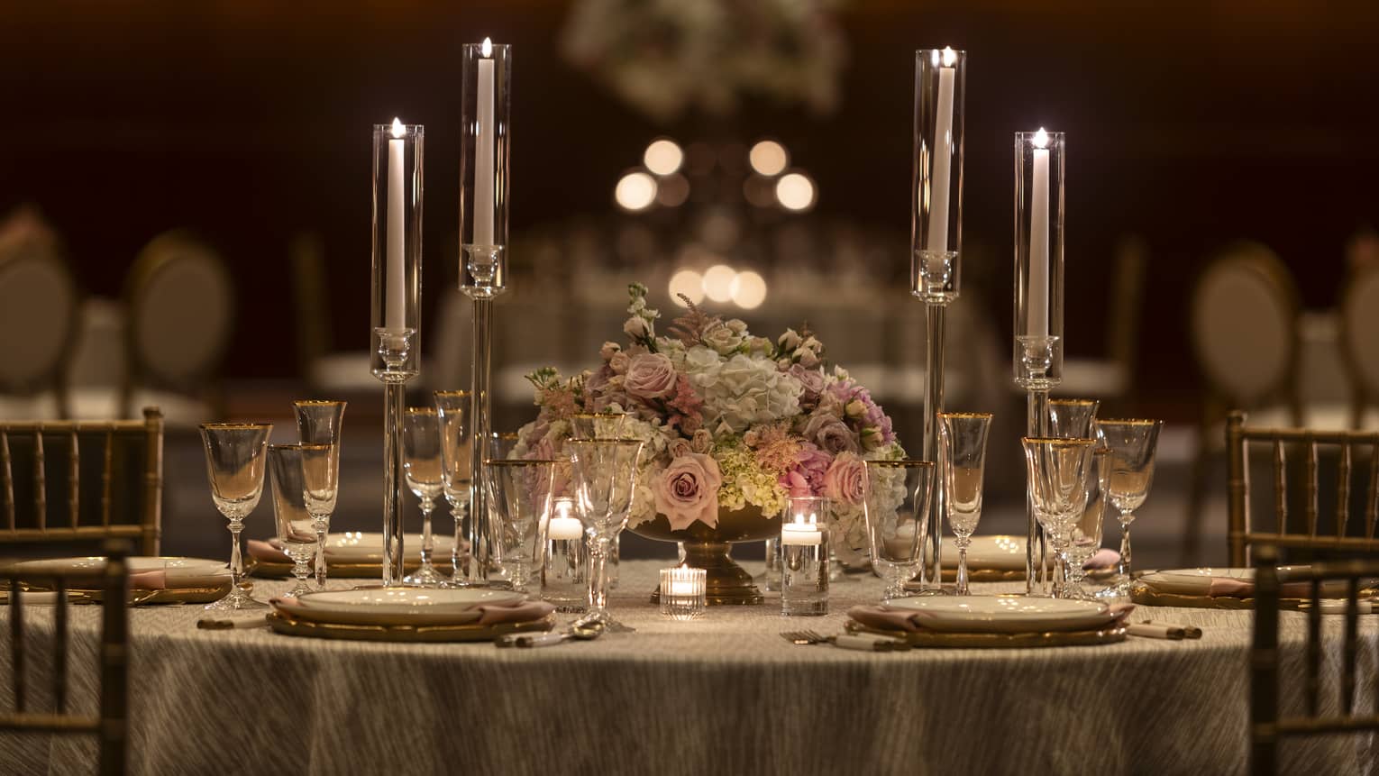 A round wedding dinner table set with glowing tealight candles in long glass holders, a colourful floral centrepiece, and tableware.