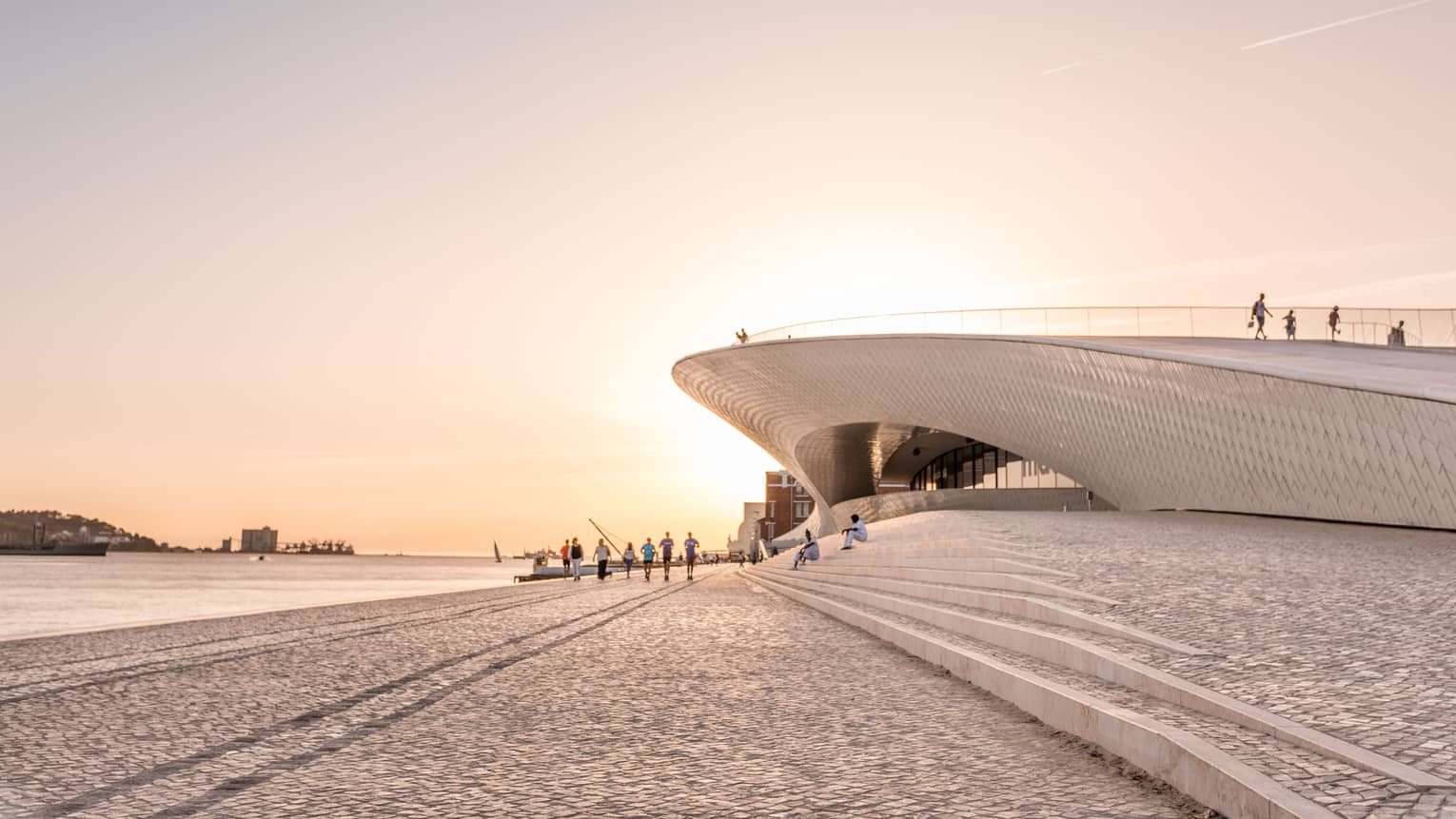Modern waterfront building with sweeping architectural curves, people walking along a cobblestone promenade at sunset.