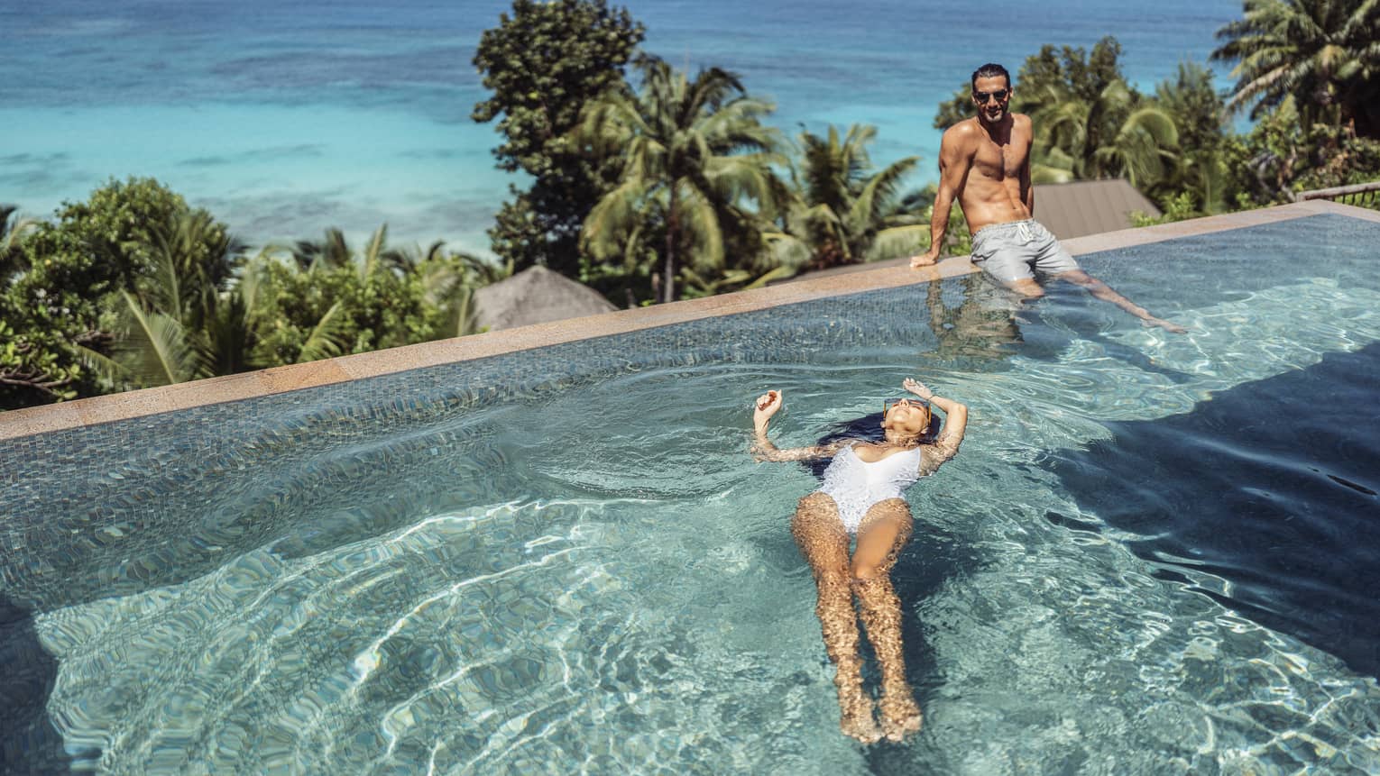 Two people in swimsuits, one floating on back in pool and one sitting on pool?s edge with palm trees and ocean in backdrop