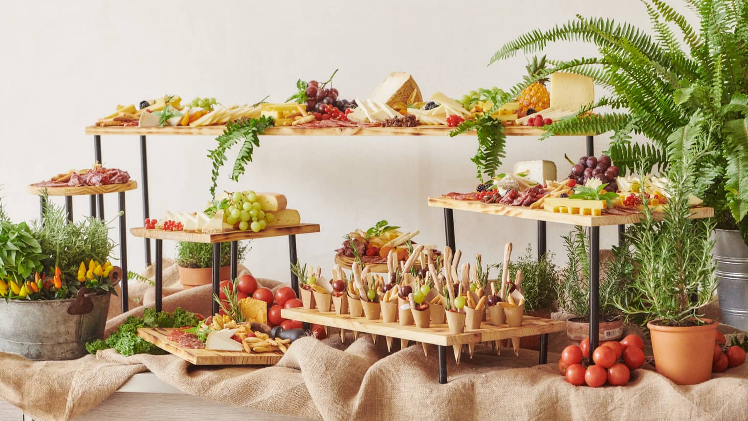 An arrangement of serving stands of various heights holding foods such as grapes and cheeses is presented on a sideboard covered in beige fabric and accented with decorative potted plants and greenery