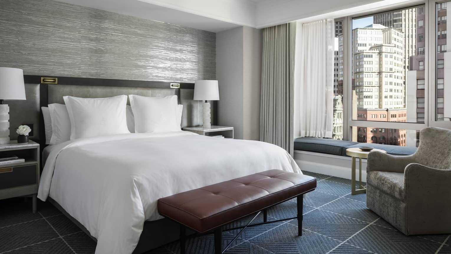 A hotel room with a bed with white sheets, a red bench, a grey chair, two white lamps, and a large window looking out at a city.