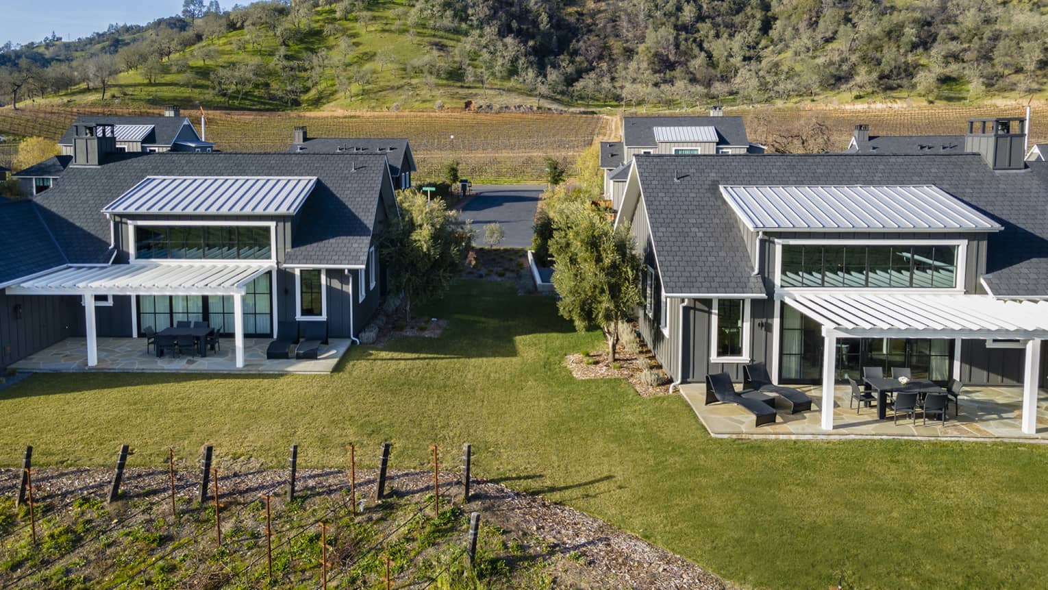Aerial view of luxury resort gray villas with patios with pergolas, surrounded by grassy lawns, vineyards and hillside scenery
