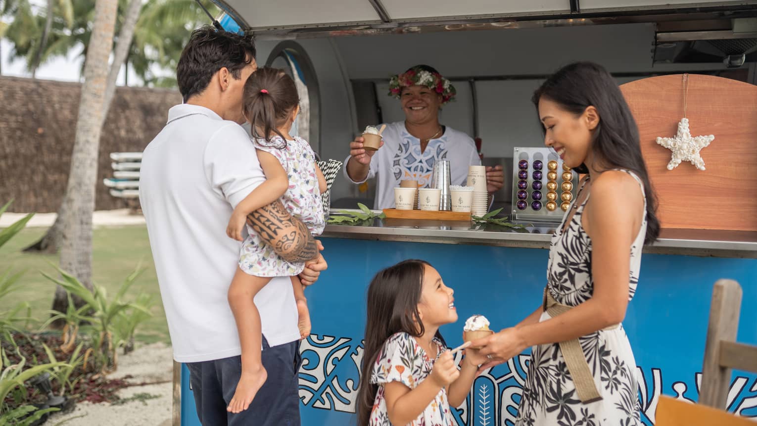 A family of four buys ice cream from a smiling employee wearing a flower crown. The stall is decorated with Polynesian art.