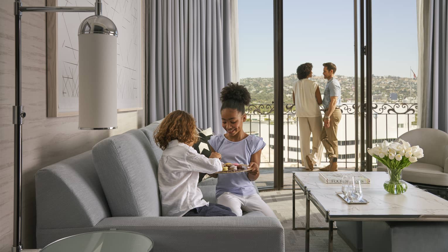 Two adults stand looking out a hotel balcony while two children play on a grey sofa inside