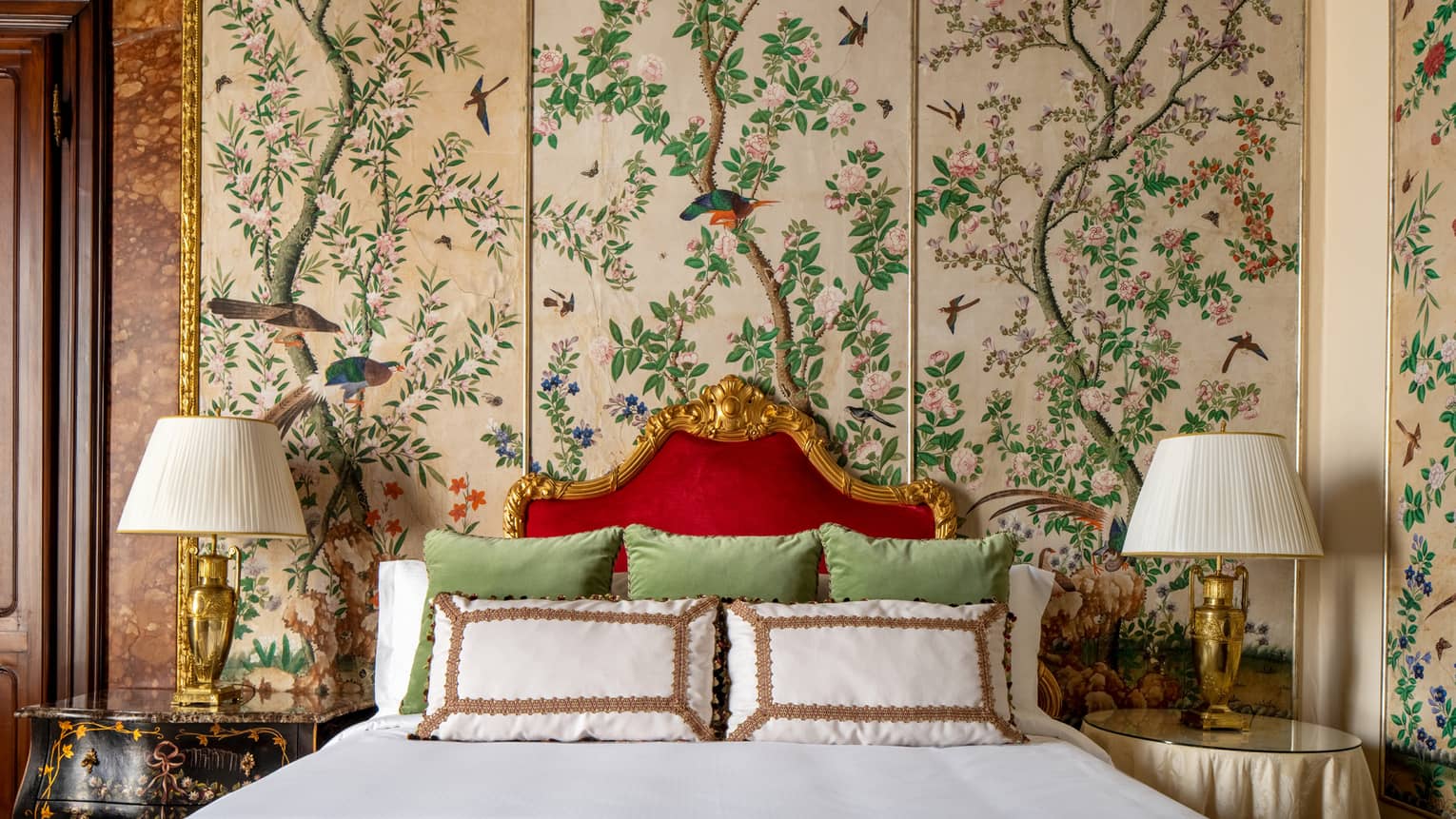 Luxurious bed with gold-framed red headboard, white pillows, and green cushions, flanked by lamps, set against a decorative wall with floral and bird patterns.