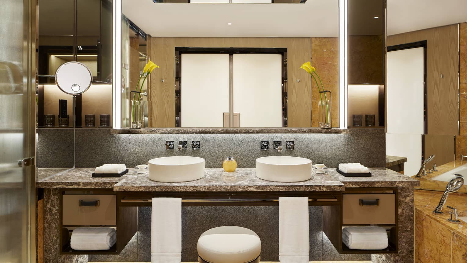 Luxurious bathroom in the Deluxe Peak-View Room with double vanity, marble countertop, tall mirrors, folded towels, a bathtub, and elegant lighting fixtures.