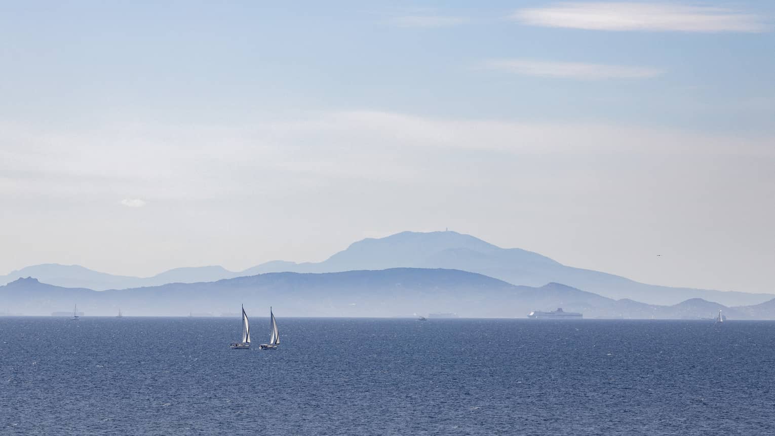 Two sailboats on blue ocean with blue-grey mountain range in the background