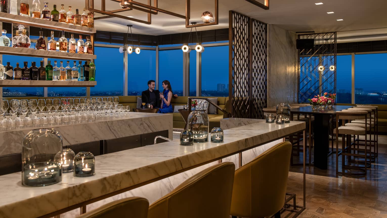 Two smiling people in formal wear sit in booth behind marble bar with liquor bottles on display