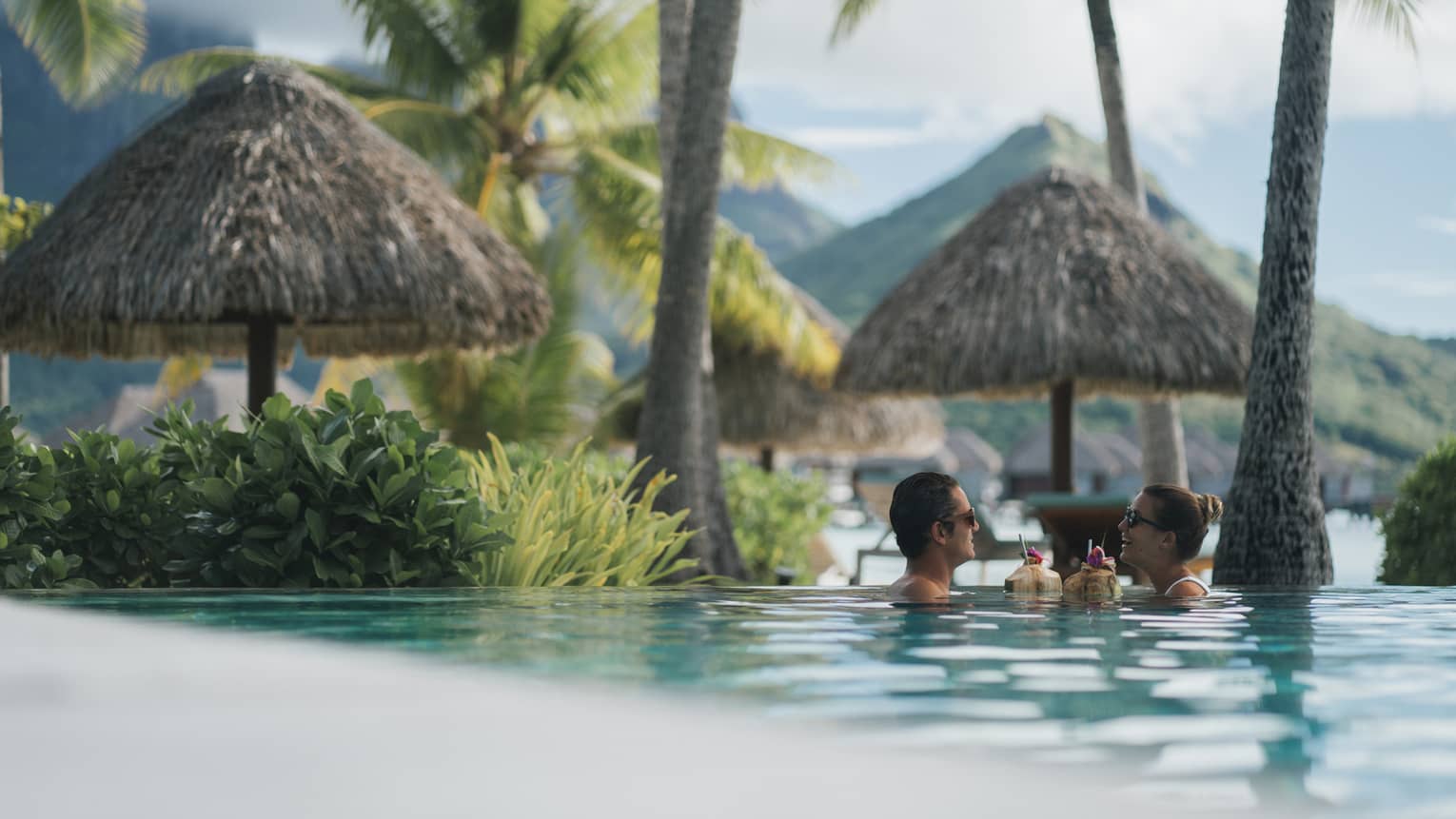 Two guests sip coconut drinks while floating in a pool surrounded by palm trees