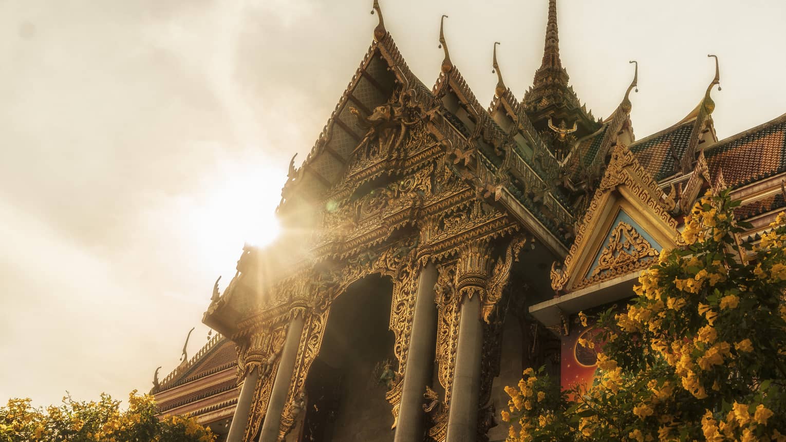 Intricately designed temple with ornate carvings and tiered roofs, illuminated by sunlight, with a tree and yellow flowers in the foreground