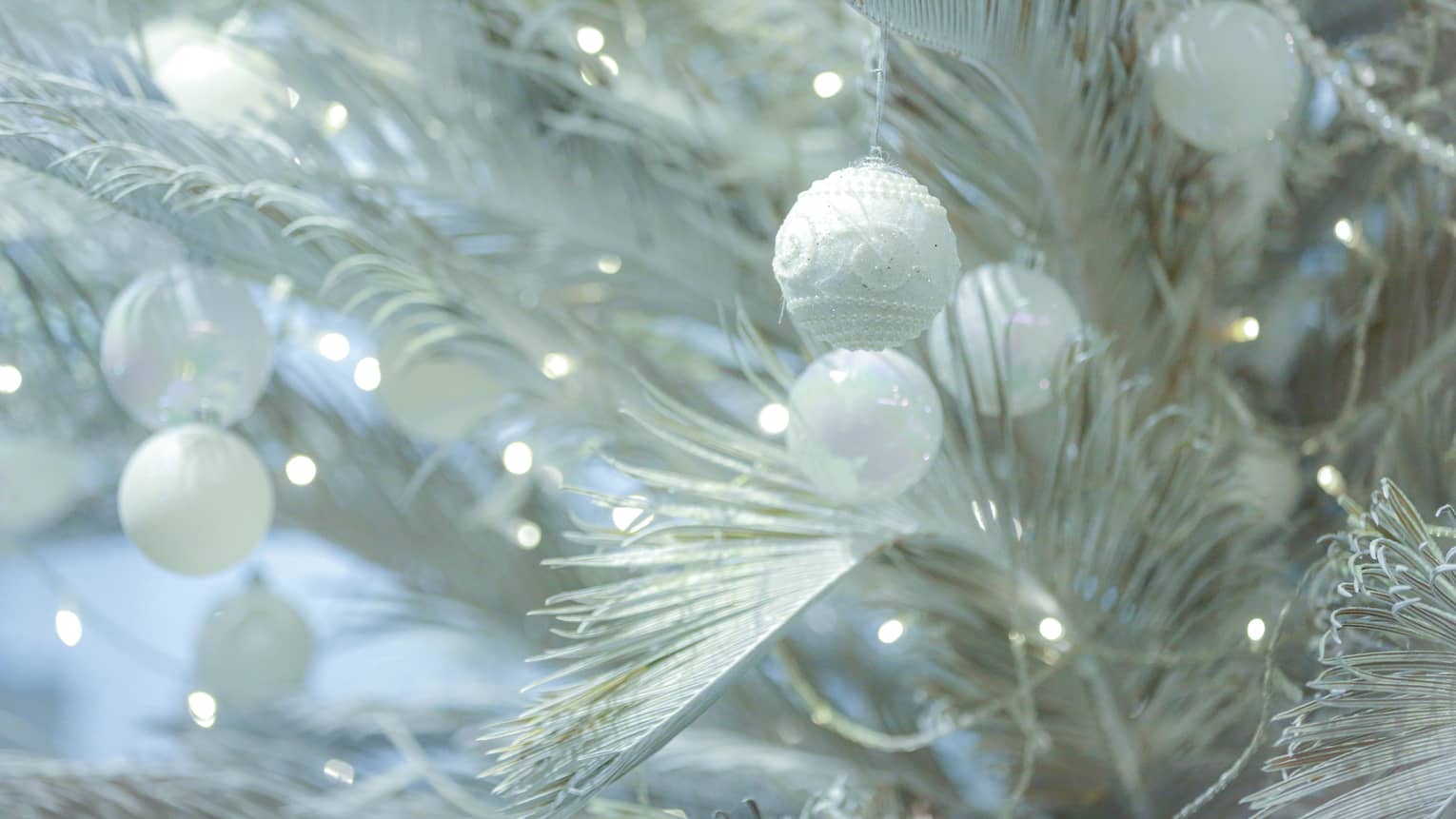 Close up of white Christmas tree branches with tiny twinkling lights and several round, white ornaments