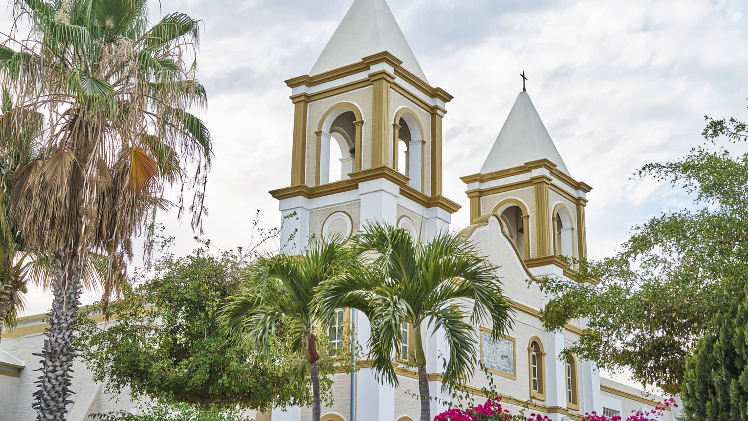 Two church bell towers rise behind palm trees and in front of a cloudy sky. They feature arched windows and gold paint.