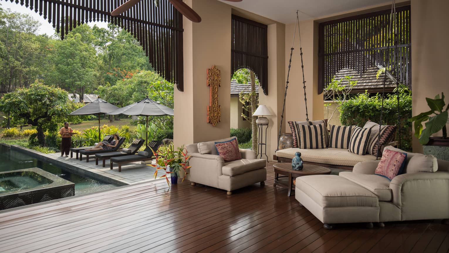 Covered outdoor lounge with cushioned seating ceiling fan and poolside view featuring sun loungers umbrellas and lush garden surroundings