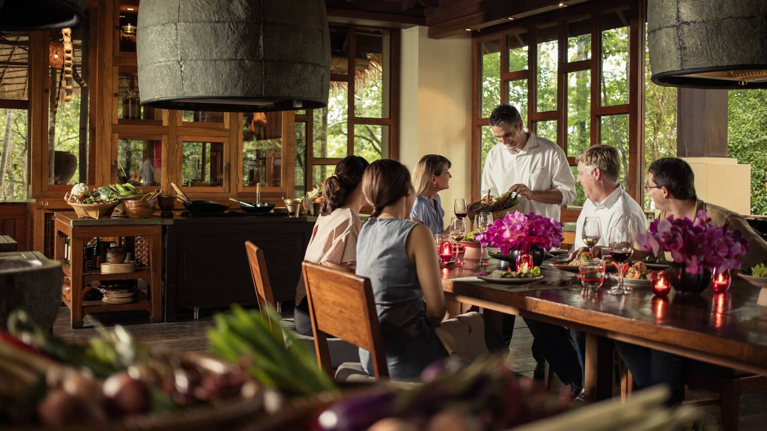 Chef presents basket of vegetables to large group of friends at table in sunny restaurant