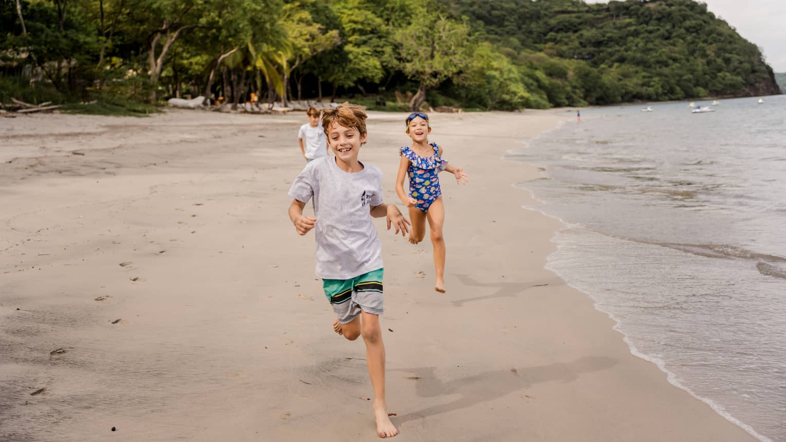 Three children run along the beach with the ocean on one side and a lush treeline behind them