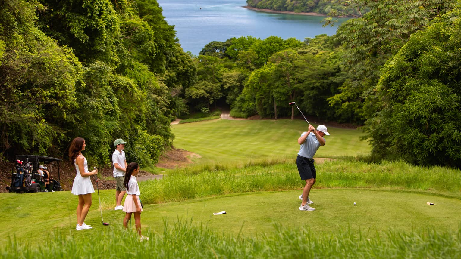 Three people stand to the side watching another person tee off onto a fairway that has the ocean in the distance