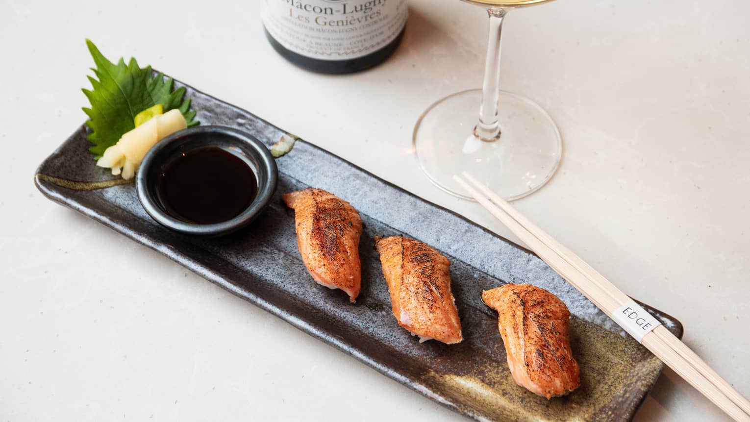 Three piece of torched salmon nigiri plated on a dark brown stoneware platter with a ramekin of soy sauce and sliced of picked ginger, with a pair of chopsticks, wine glass and a bottle of wine on the side