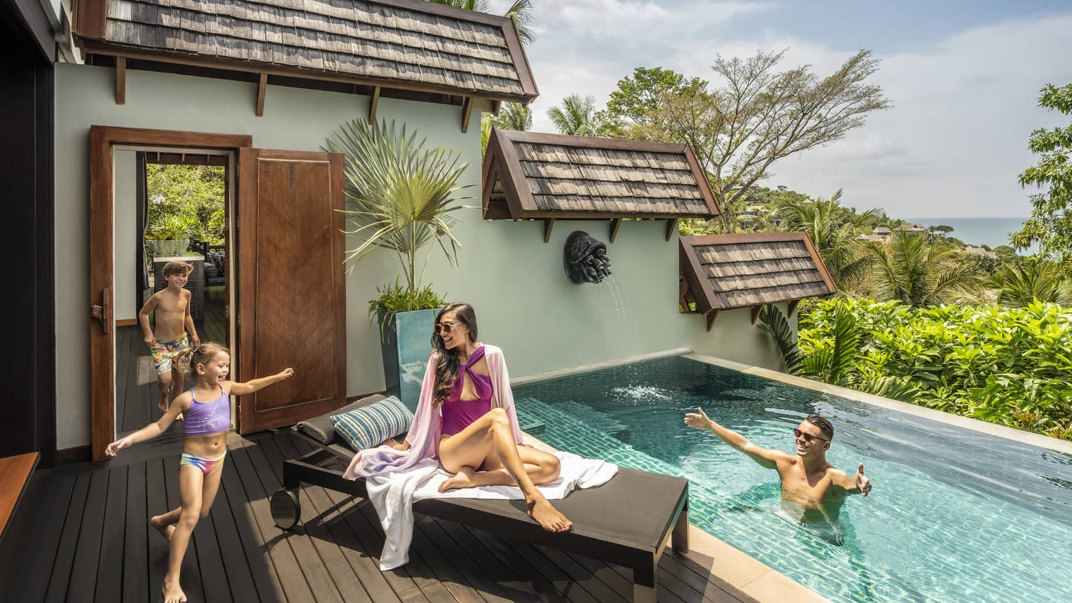 Two young children run into a small pool. Woman sits on lounge chair and man welcomes children in the pool.