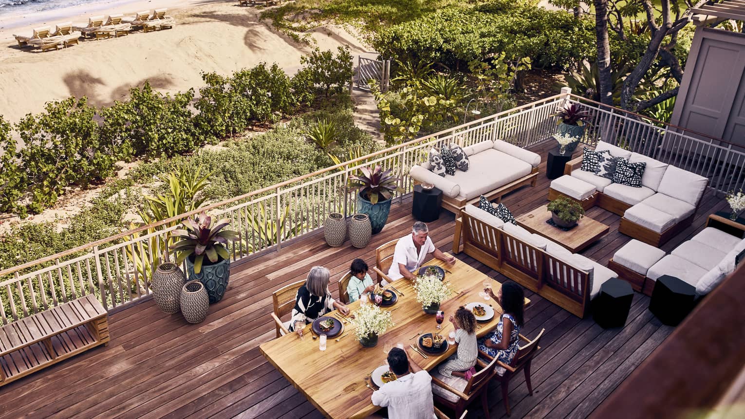 A view from above of a family enjoying a meal together on a deck with ample sitting and merely steps away from a small beach.