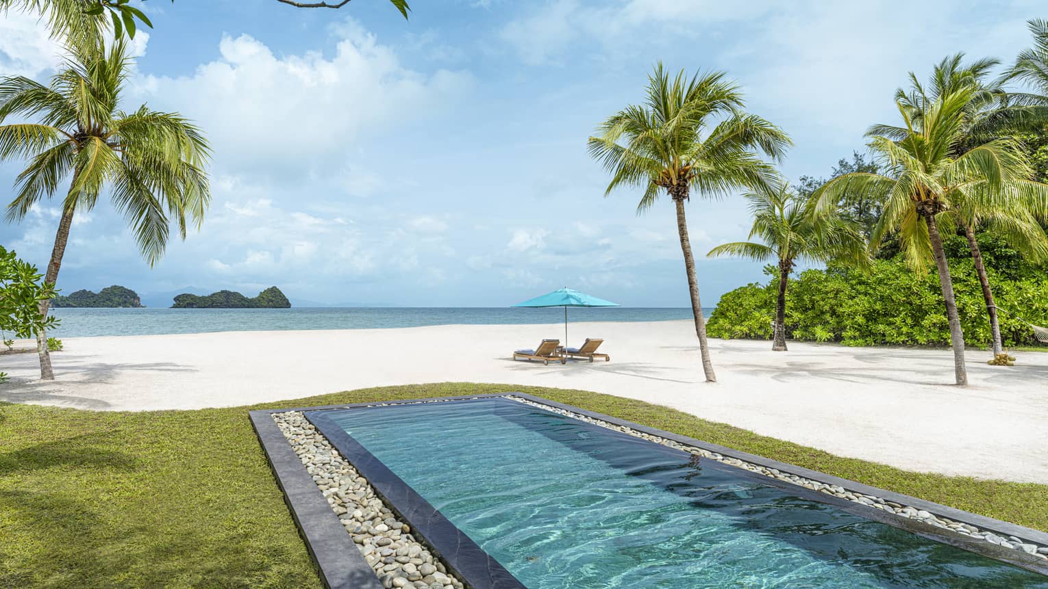 Rectangular in-ground pool jutting out to white-sand beach dotted with palm trees with two lounge chairs and umbrella along blue waters