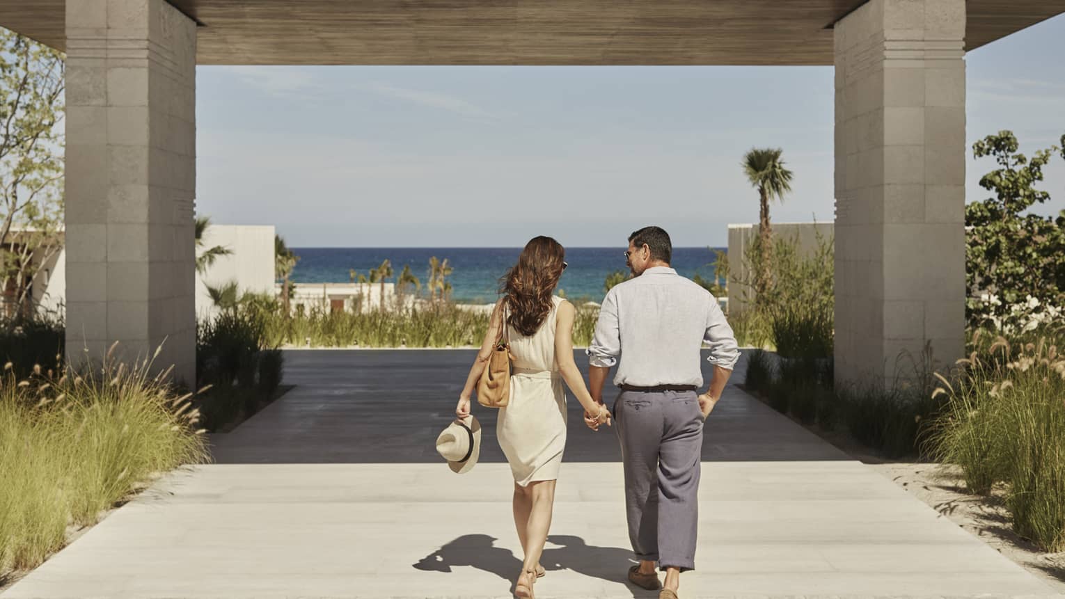 Rear view of a couple walking hand in hand towards a gradient blue ocean on a covered boardwalk lined by tall grasses.