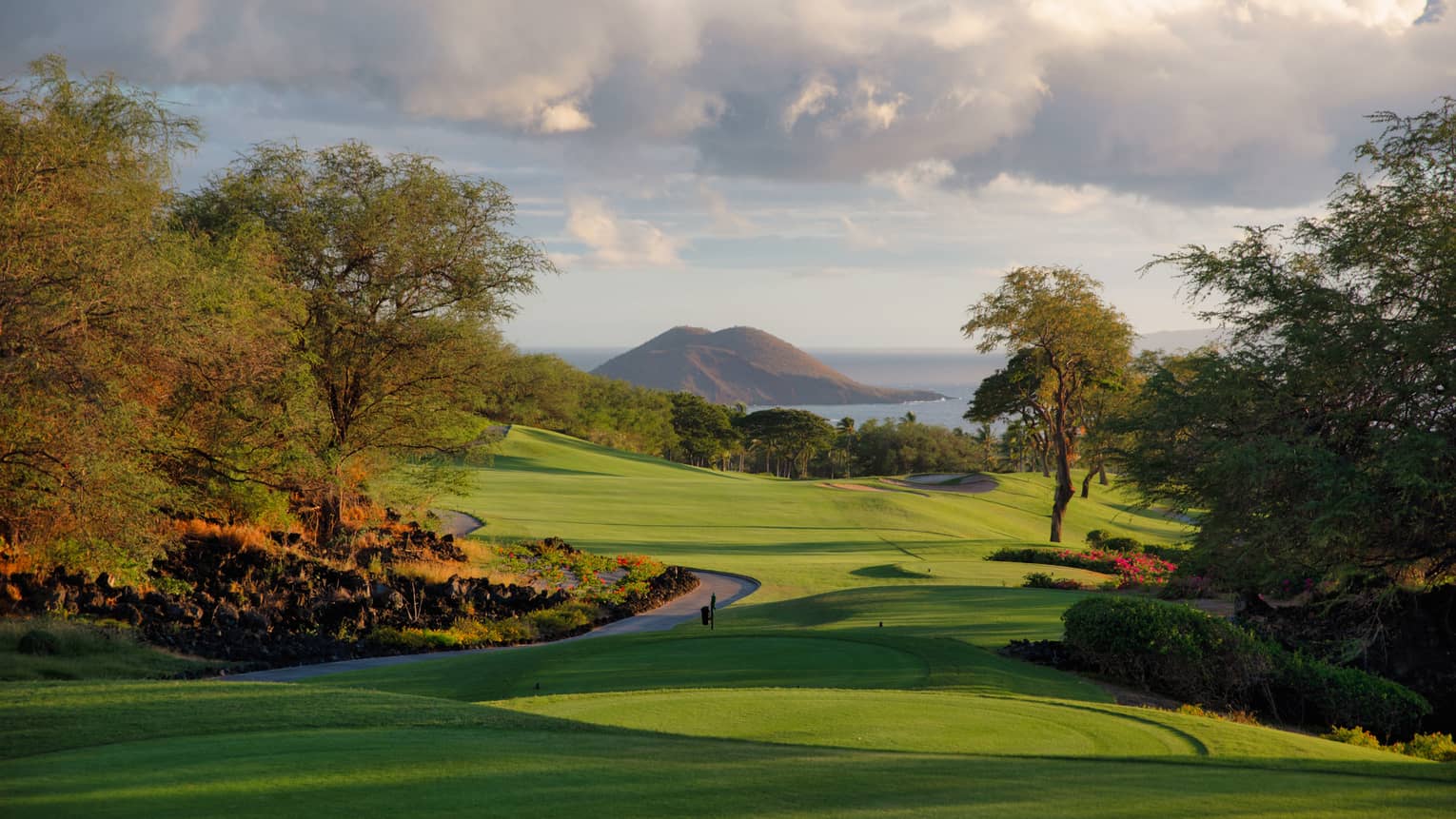 Long shadows stretch over a bright golf course bordered by lush trees and expansive water under fluffy, low-hanging clouds.