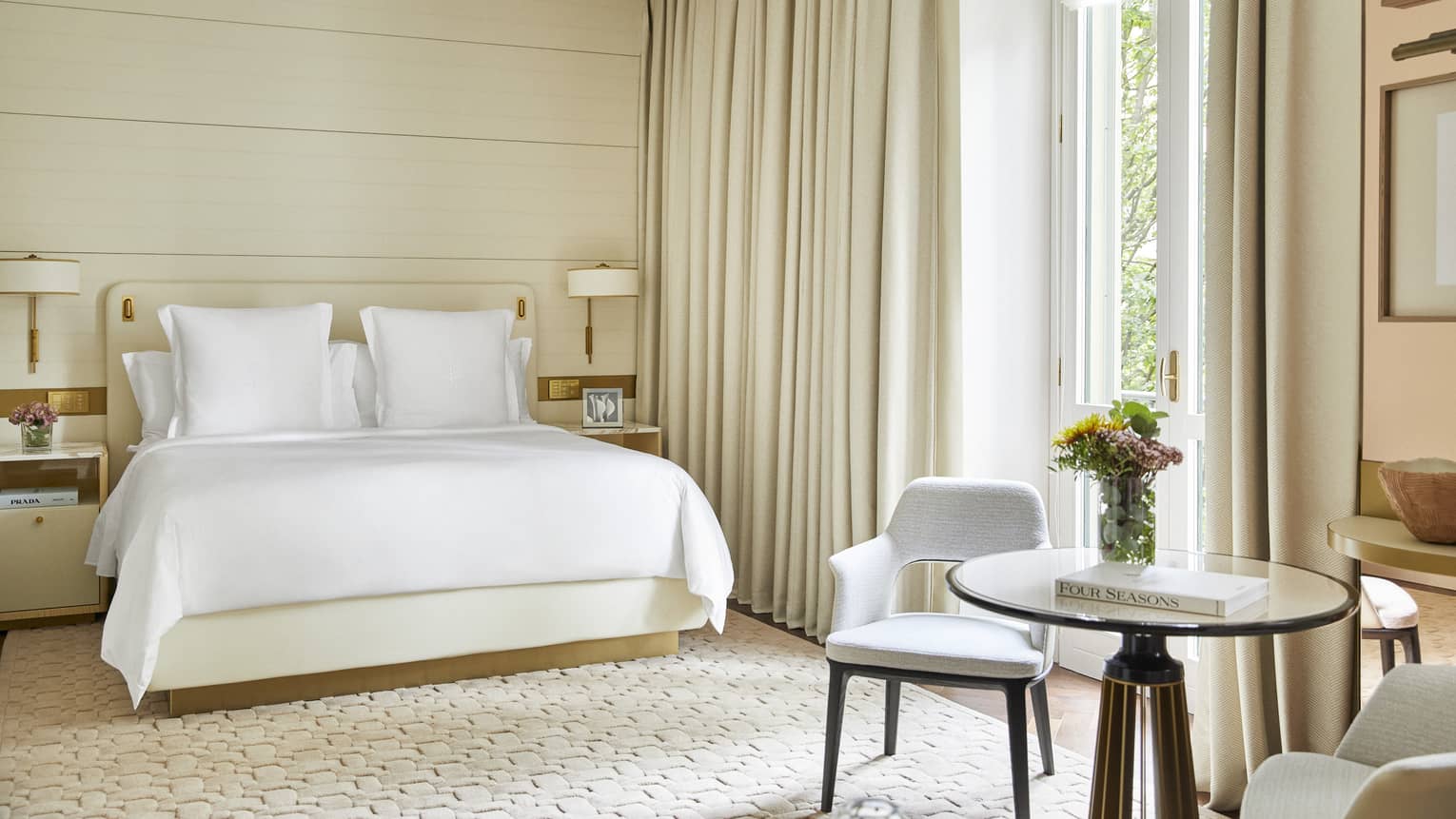 Light-filled hotel guest room with king bed, small round table with two chairs and creme-coloured curtains