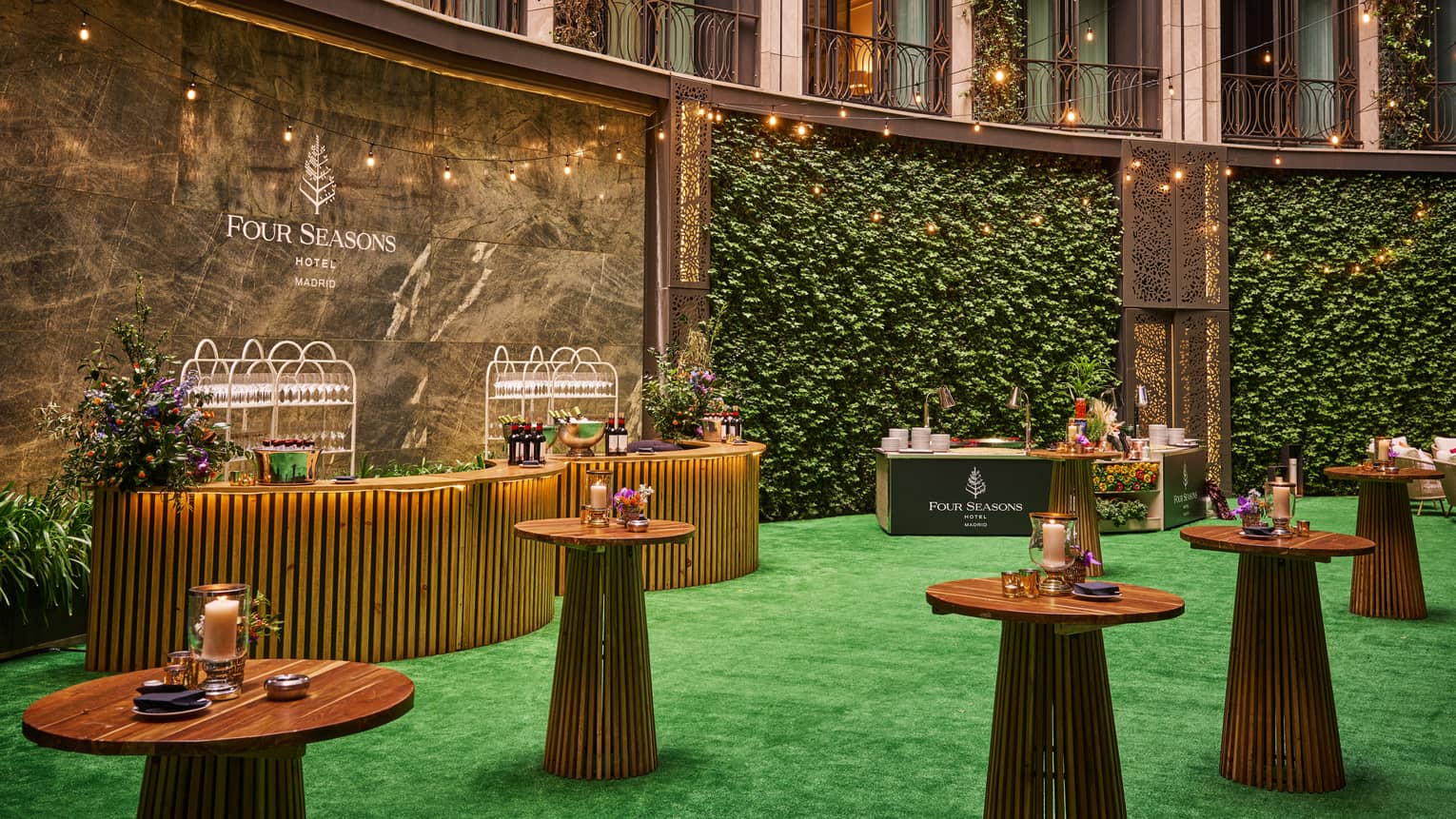 Interior courtyard with green turf flooring and vine-covered walls set up for an event with wooden cocktail tables and multiple display tables