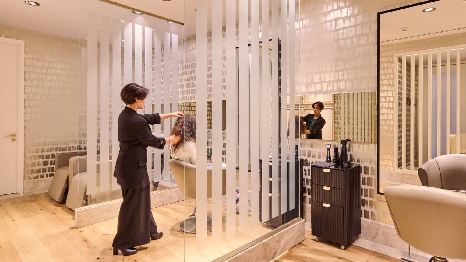 A hair stylist styles a guest?s hair in a brightly lit salon with pale wood floors, large mirrors, salon chairs and contemporary decor.