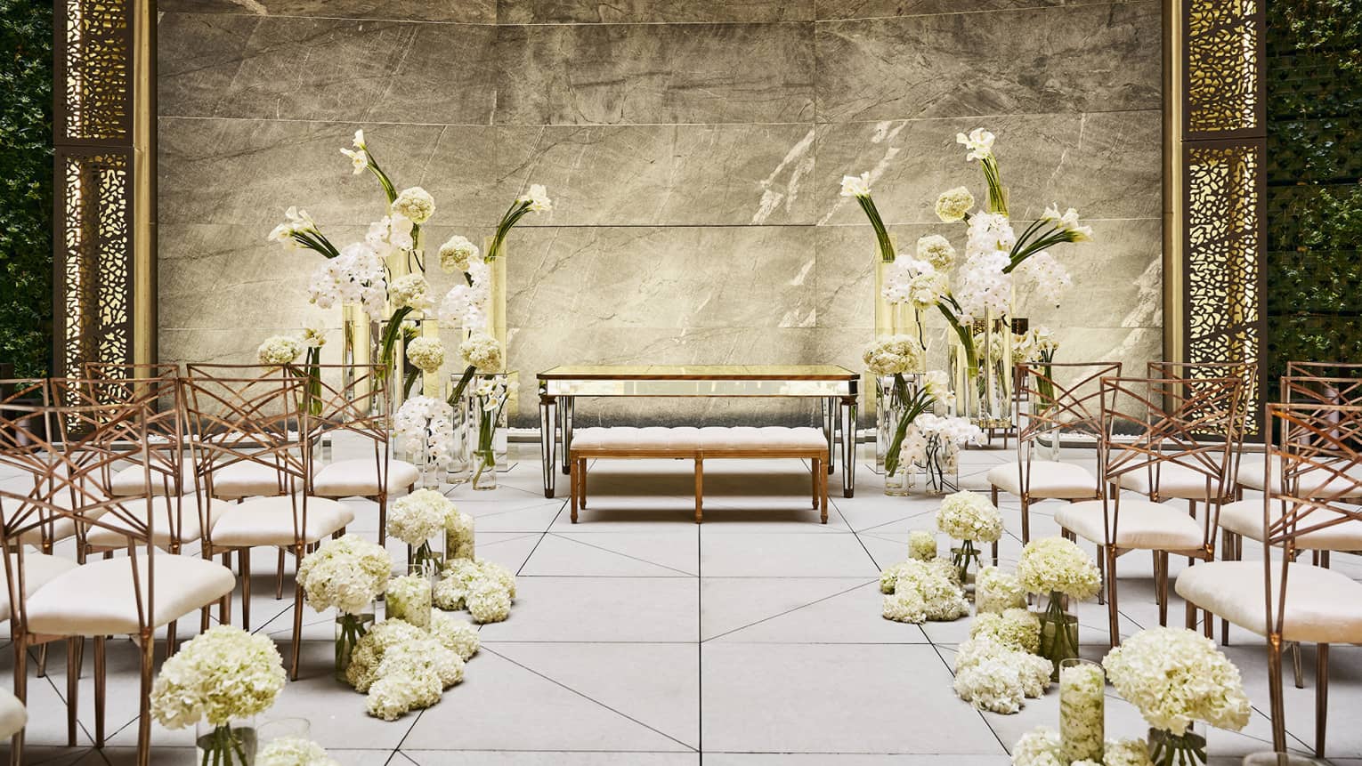 An elegant indoor wedding ceremony setup with white floral arrangements, modern chairs and a marble backdrop, featuring intricate gold latticework.