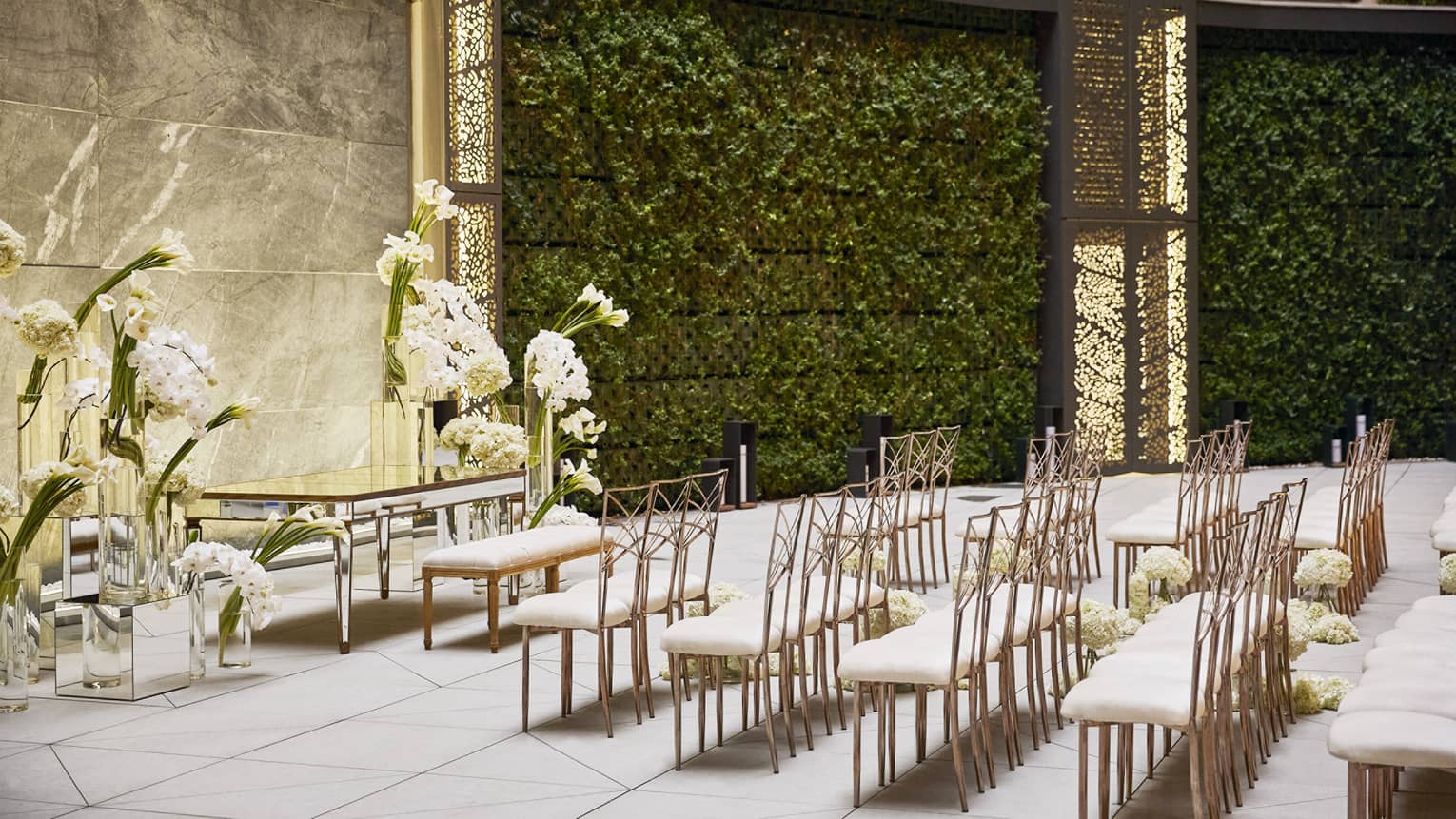 Five rows of white-and-gold chairs set up facing an antique table and bench surrounded by large white floral arrangements, all set in an intertior courtyard featuring curved, vine-filled walls