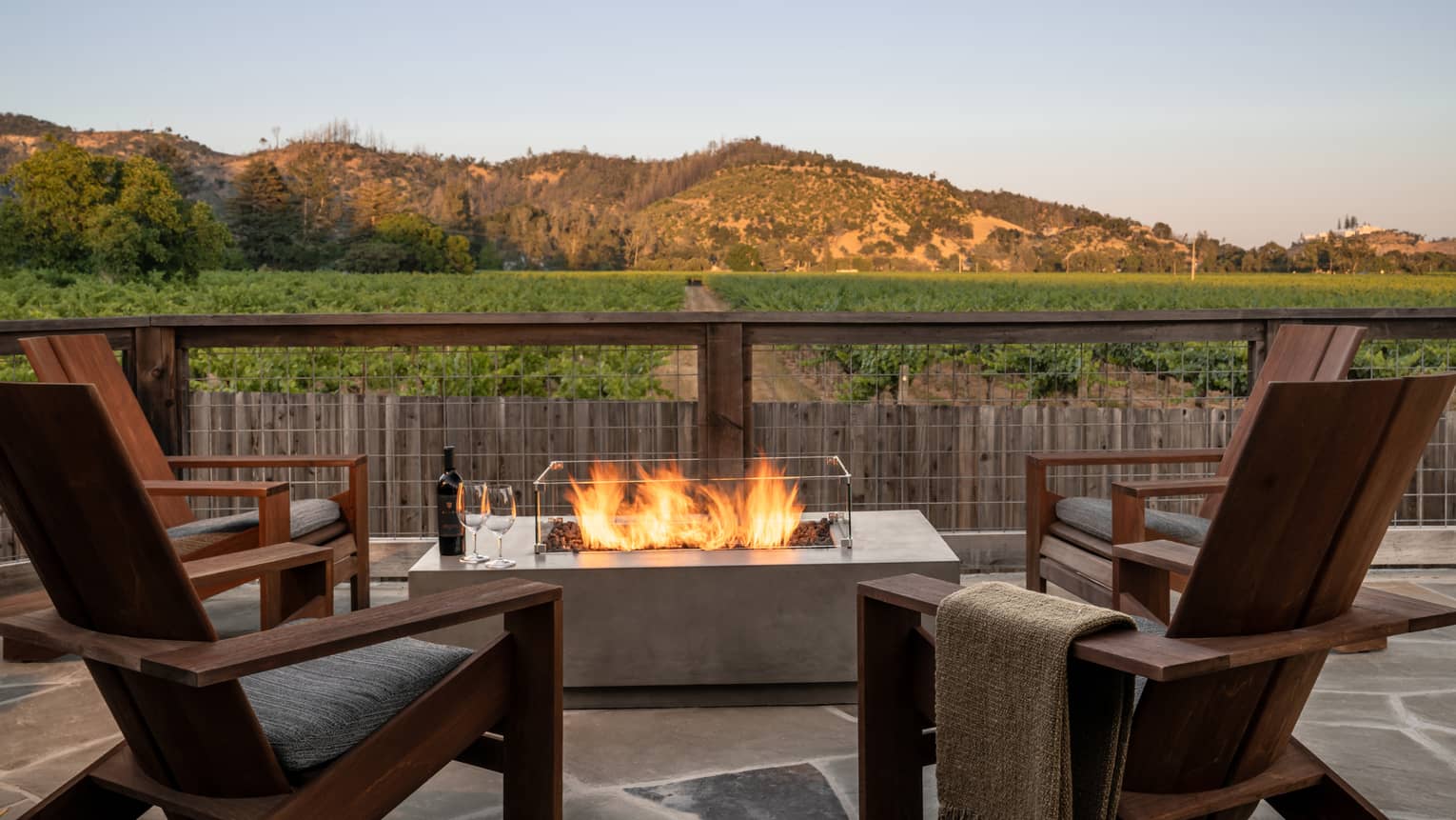 Four wooden chairs surround a bonfire on a stone terrace that overlooks vineyards