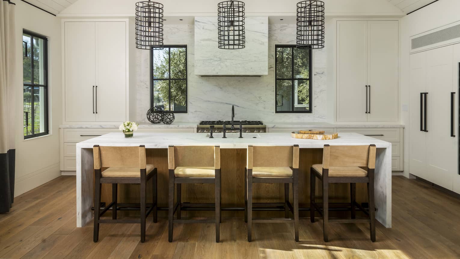 Luxury resort kitchen with marble island, four beige barstools, black pendant lights, large white cabinets, stove and three windows overlooking greenery