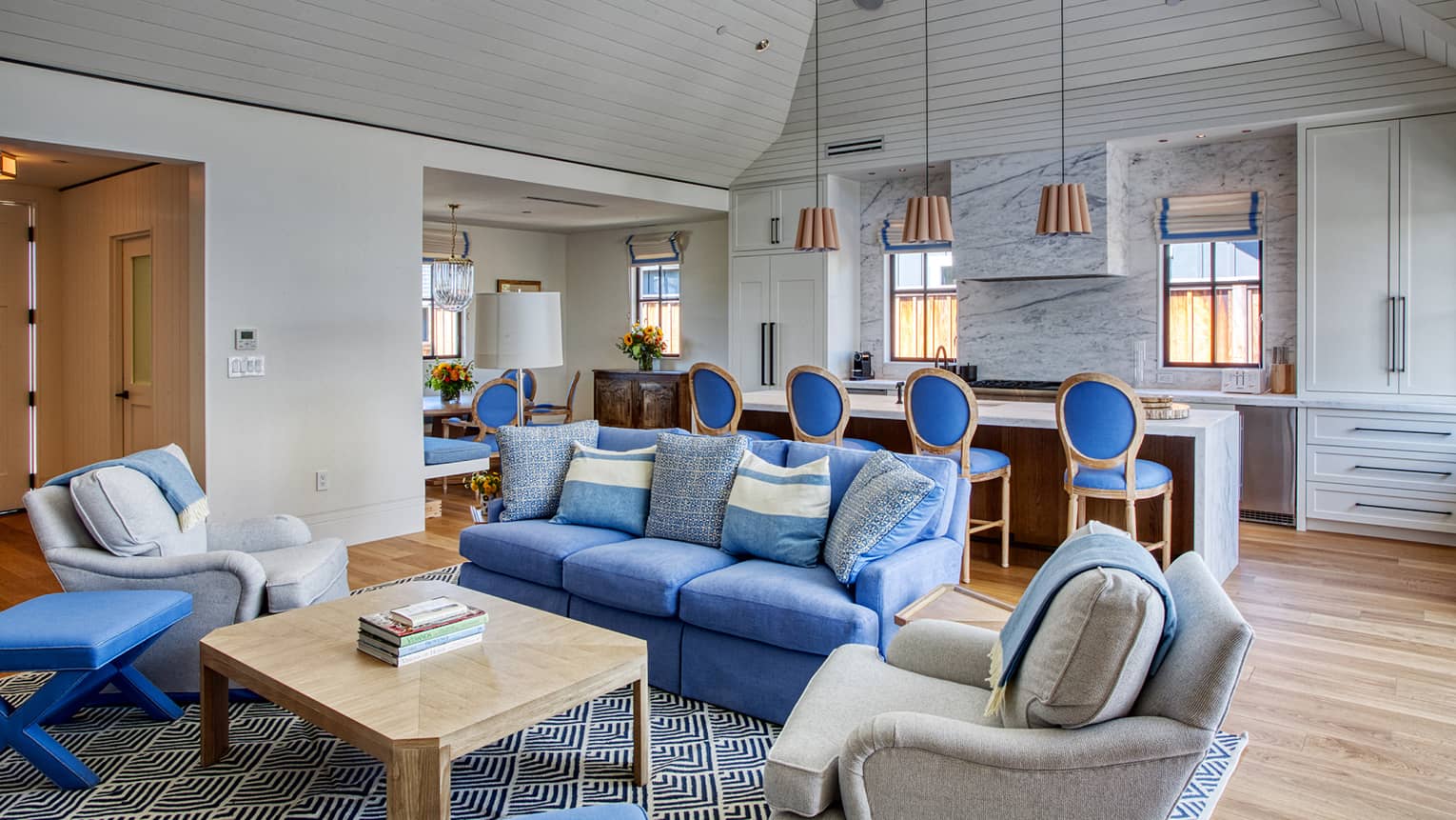 Luxury resort living room and kitchen area with a blue couch, grey linen chairs, wooden coffee table and large island with pendant lights