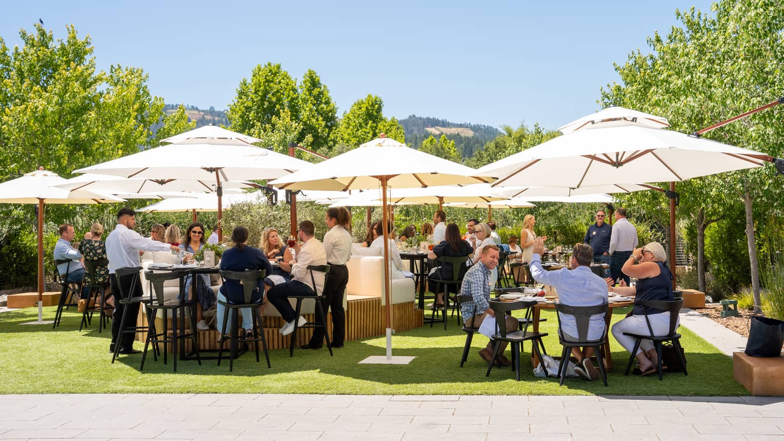 People sitting at tables with umbrellas on a lawn surrounded by trees