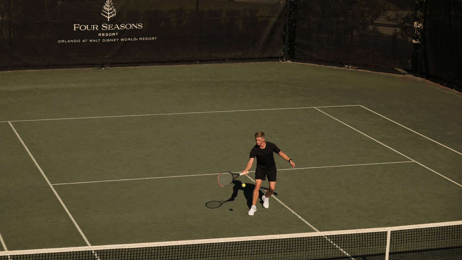 Guest wearing black shirt and shorts swings tennis racket to hit the ball on a tennis court