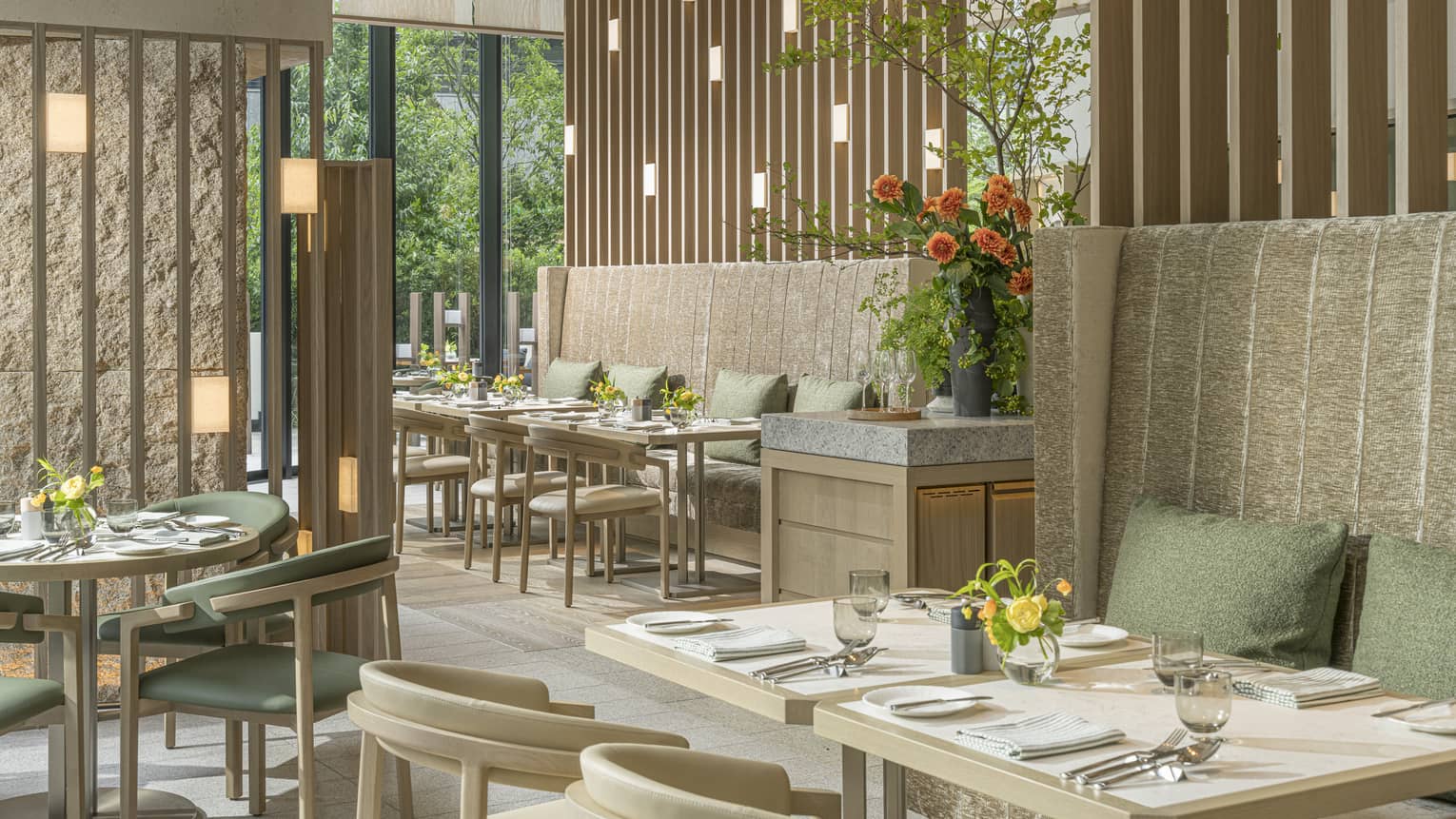 Airy earth-toned bistro with green cushions on tall banquettes, vases of flowers on tables and windows showing lush foliage.