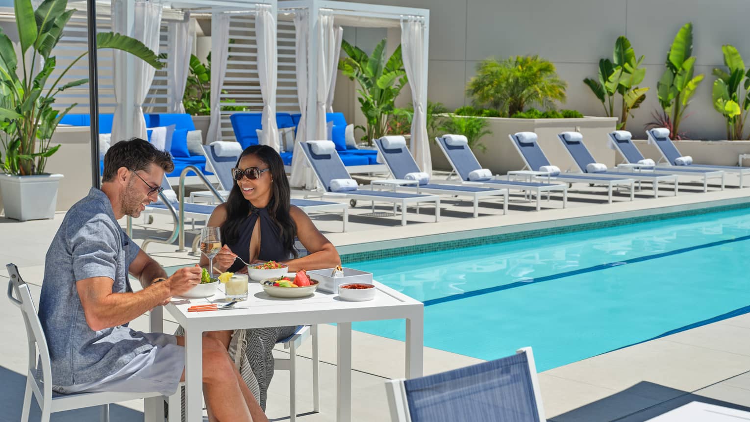 Two guests sitting at a table under a blue umbrella by the pool.