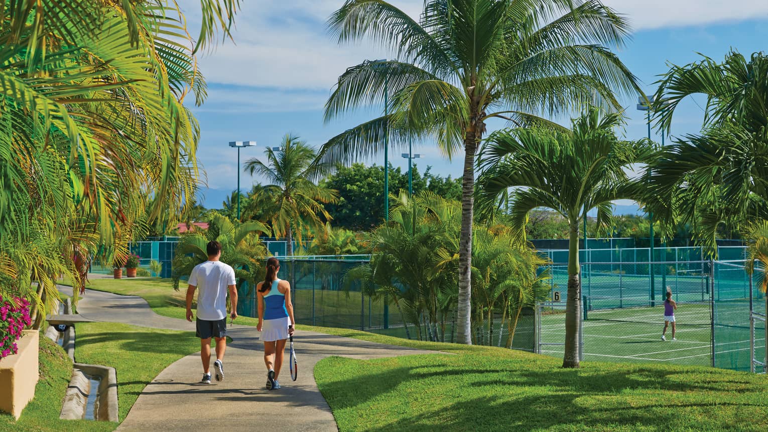 A man and woman walking along a path next to tennis courts and palm trees.