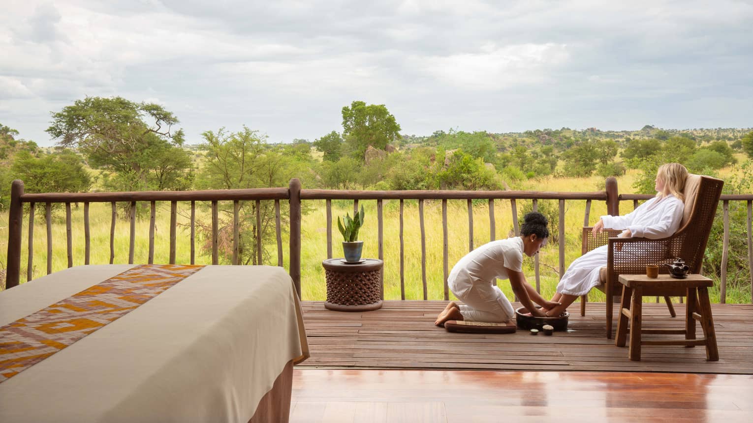 Spa therapist bathing fee of seated robed guest on spa terrace, looking out to expansive plain over vertical wooden railing