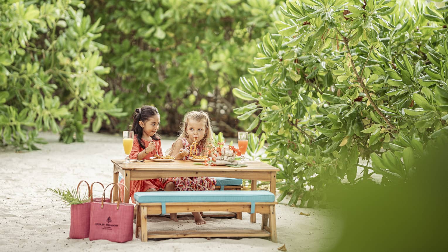 Smiling children enjoy a meal at a kid’s picnic table with cushioned seats on a sandy area surrounded by vegetation.