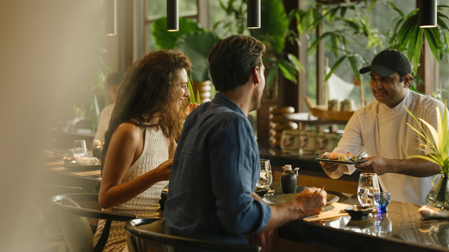 A smiling couple sit at a bar top in a restaurant decorated with vibrant plants as a chef hands them a sushi platter.