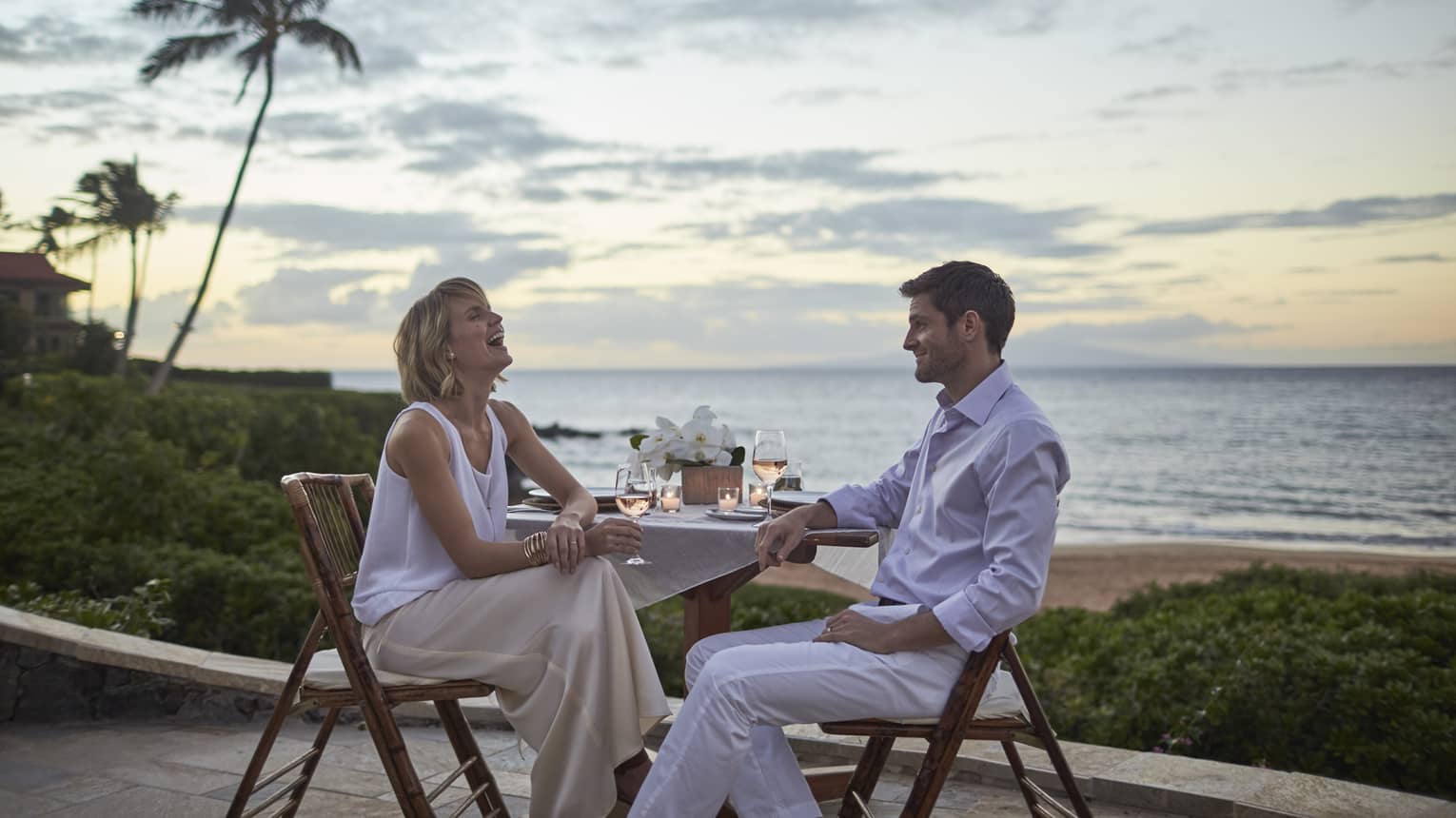 Two people enjoying a beachfront dinner at sunset with wine and ocean view