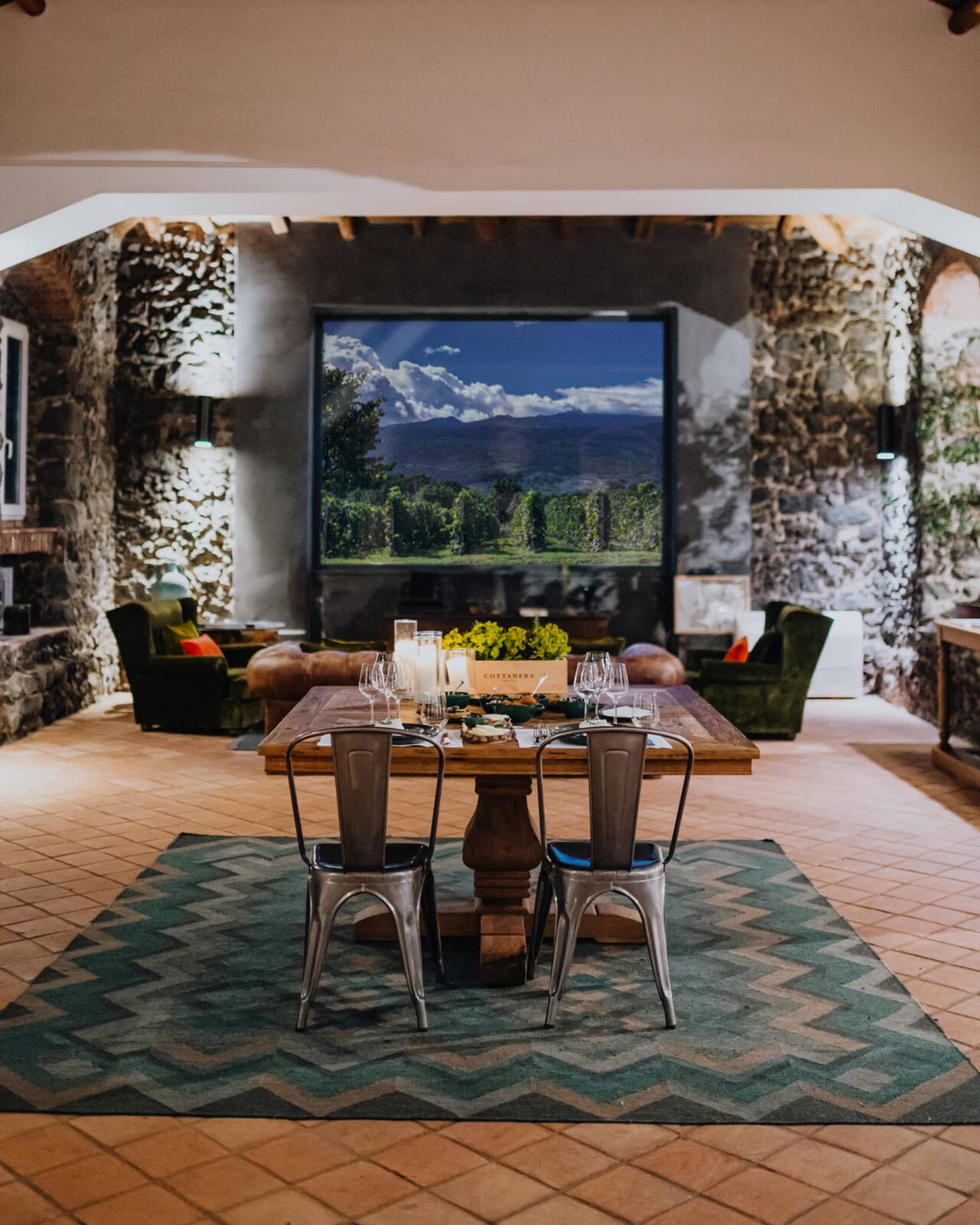 A wooden dining table set with floral arrangements and glassware, in a room with a window overlooking mountains and a vineyard