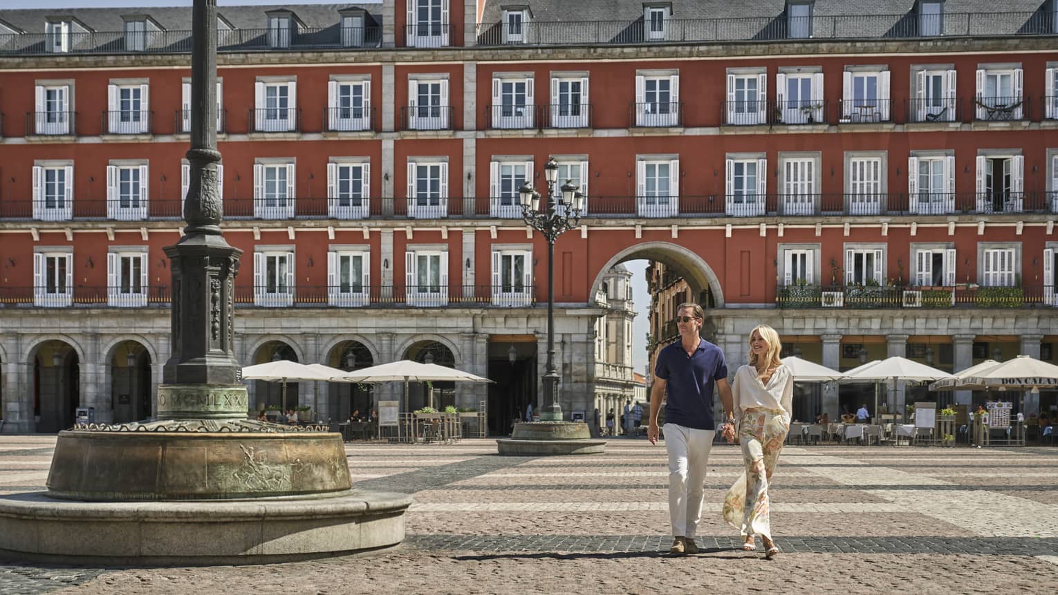 A man and woman walking along a stone paved street with a large building behind them.